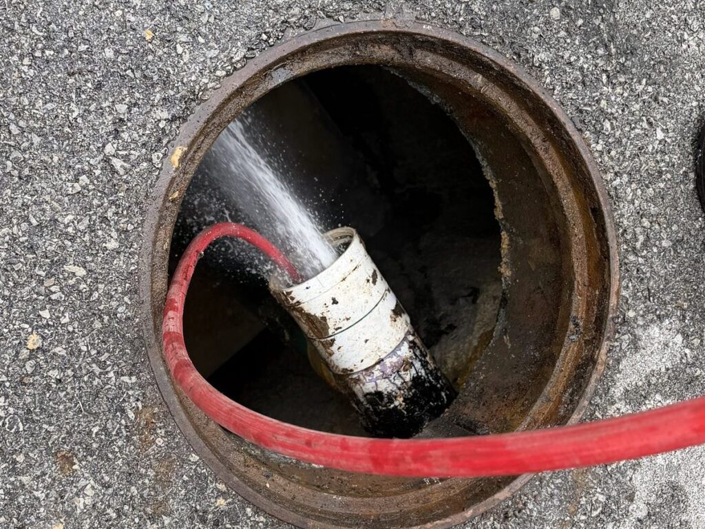 Hydro jetting a sewer line: high-pressure water sprays from the pipe inside a manhole as the red jetting hose runs into the line